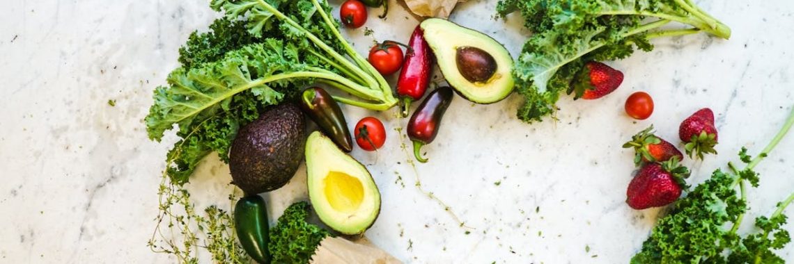 Avocado, kale, and more fresh produce artfully arranged on a marble surface.